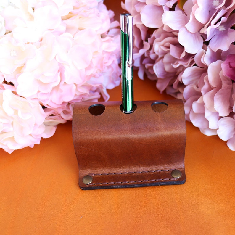 May include: A brown leather pen holder with three slots. A green pen is in one of the slots.
