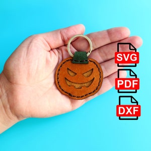 May include: A brown leather pumpkin keyring with a stitched outline and a cut-out jack-o'-lantern face. The keyring is attached to a gold metal ring and a green leather loop. The image also shows the file formats available for download: SVG, PDF, and DXF.
