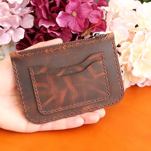 May include: A brown leather wallet with a zipper closure and two interior pockets. The wallet has a distressed look and is made of high-quality leather.