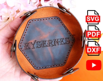 May include: A round, brown leather tray with a dark brown, hexagonal centre. The tray is embossed with the word "EYSERNES". The tray has metal snap closures and is designed to hold small items. The tray is shown against an orange background.