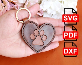 May include: A brown leather heart-shaped keychain with a paw print design. The keychain has a gold-colored ring and is stitched around the edges. The background includes flowers and file type icons for SVG, PDF, and DXF.