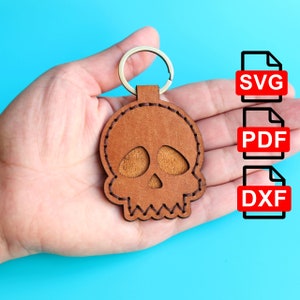 Può includere: Un portachiavi in ​​cuoio marrone a forma di teschio con un anello portachiavi argentato. Il teschio presenta dettagli cuciti e un contorno cucito. Il portachiavi è tenuto in una mano su uno sfondo blu. L'immagine include anche tre icone di tipo di file: SVG, PDF e DXF.