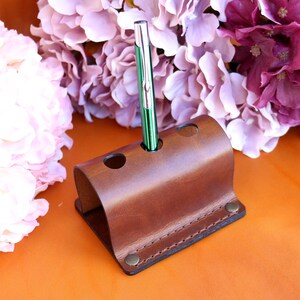 May include: A brown leather pen holder with three slots. A green and silver pen is in one of the slots.