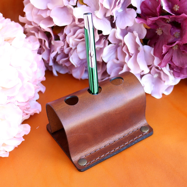 May include: A brown leather pen holder with three slots. A green and silver pen is in one of the slots.