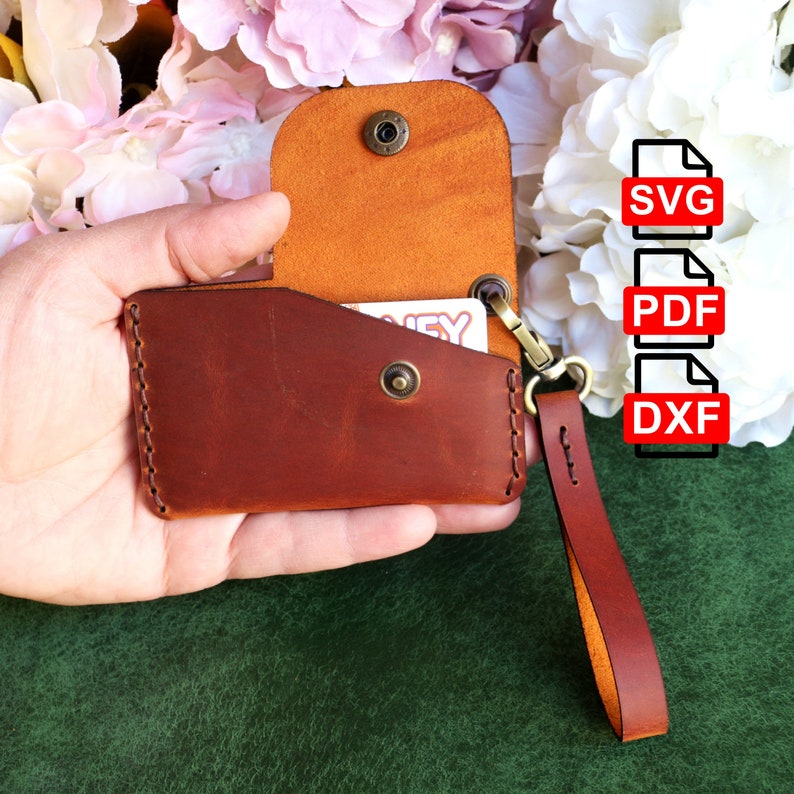 May include: A brown leather card holder with a flap and a snap closure. The holder has a small strap with a metal loop. The holder is open and a card is visible inside. The holder is being held in a hand.
