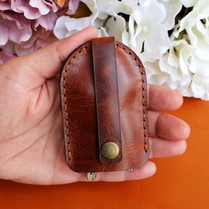 May include: A brown leather key holder with a flap closure and a metal button. The key holder is held in a hand.