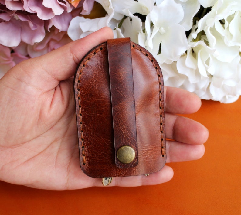 May include: A brown leather key holder with a flap closure and a metal button. The key holder is held in a hand.