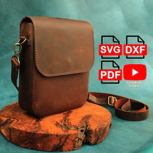 Puede incluir: Un bolso bandolera de cuero marrón con cierre de solapa y correa ajustable. La bolsa se muestra sobre una superficie de madera. La imagen también incluye iconos con el texto "SVG", "DXF", "PDF" y "Tutorial Video".