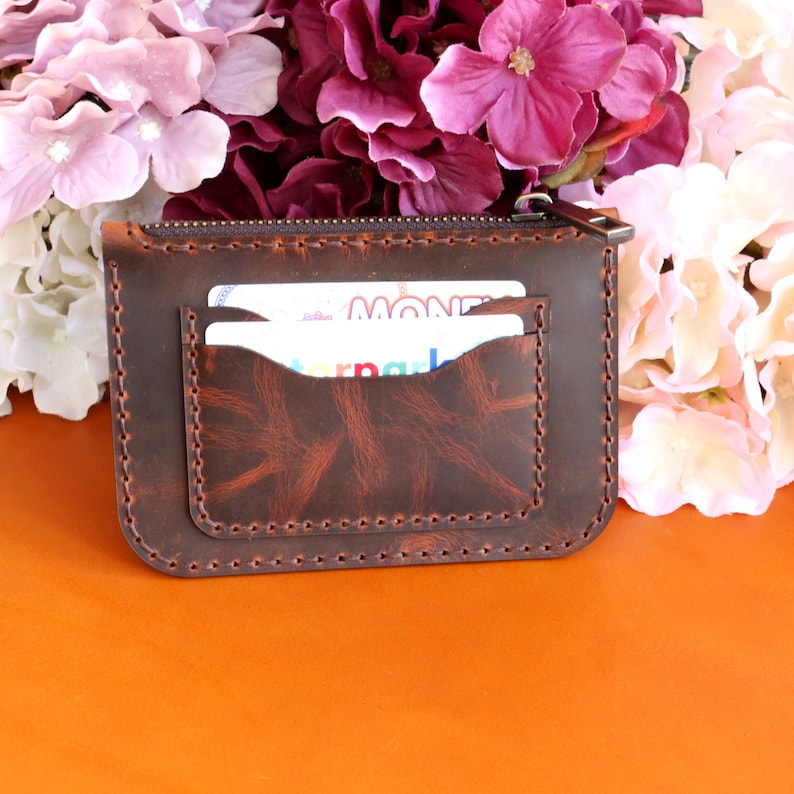 May include: A brown leather zippered wallet with two card slots and a stitched design. The wallet is lying on an orange surface.
