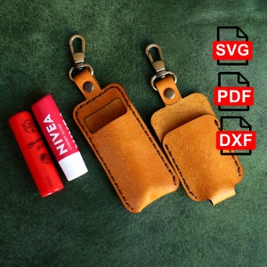 May include: Two brown leather keychains with a metal loop and a stitched pocket for a lip balm. The keychains are on a green surface with two lip balms, one red and one white with the text "NIVEA CHERRY SHINE". The image also includes text for SVG, PDF, and DXF file formats.