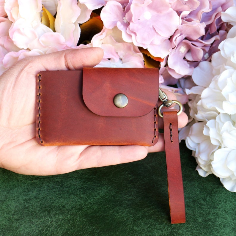 May include: A brown leather wallet with a flap closure and a small pocket on the front. It has a metal clasp and a brown leather strap attached.