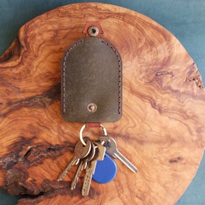 May include: A green leather key holder with a stitched edge and a metal snap closure. A set of keys with a blue fob is attached to the holder.