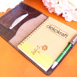 May include: A brown leather notebook cover with a spiral bound notebook inside. The notebook has a tan cover with the text "dekokraft" printed on it. A green pen is tucked into a leather pen holder on the cover.