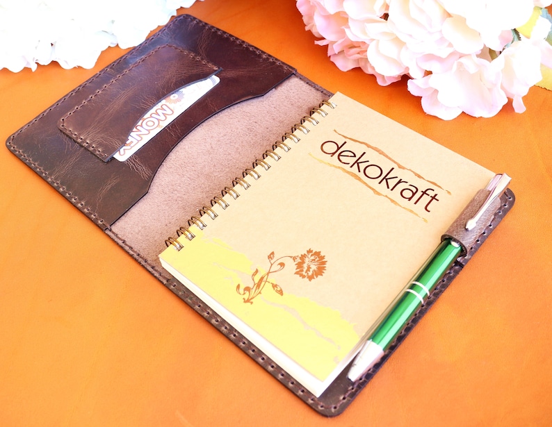 May include: A brown leather notebook cover with a spiral bound notebook inside. The notebook has a tan cover with the text "dekokraft" printed on it. A green pen is tucked into a leather pen holder on the cover.