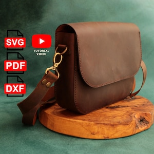 Puede incluir: Un bolso bandolera de cuero marrón con solapa y correa ajustable. El bolso tiene herrajes dorados y se muestra sobre una superficie de madera. La imagen también incluye iconos para archivos SVG, PDF y DXF, y un vídeo tutorial de YouTube.