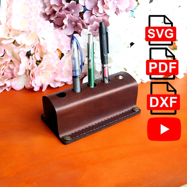 Op de afbeelding: Bruine leren pennenhouder met vier vakken voor pennen en potloden. De houder staat op een fel oranje oppervlak. De afbeelding bevat ook een set pictogrammen voor SVG, PDF, DXF-bestandsformaten en een afspeelknop.