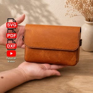 Può includere: Una borsa rettangolare in pelle color cuoio con patta ripiegabile, cuciture scure lungo i bordi e anelli laterali in metallo. L'immagine include etichette di testo per i formati SVG, PDF, DXF e un'icona di riproduzione rossa Tutorial Video.