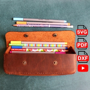 May include: A brown leather pencil case with a snap closure, open to reveal coloured pencils and pens. Several pens and a pencil are outside the case. The case is on a teal surface.