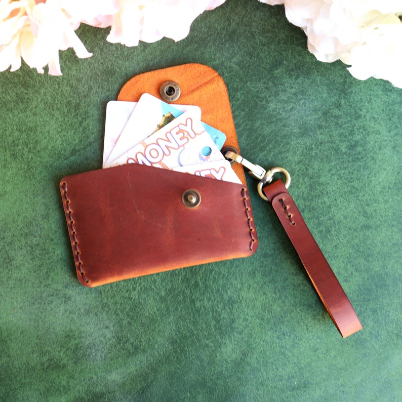 May include: A brown leather wallet with a strap and a snap closure. The wallet is open and has several cards inside. The wallet is on a green surface.
