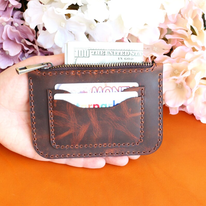 May include: A brown leather wallet with a zipper closure and two card slots. The wallet is held in a hand and has a distressed look.