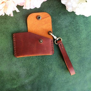 May include: A brown leather wallet with a fold-over flap and a snap closure. The wallet has a brown leather strap with a metal clasp. The wallet is on a green background.