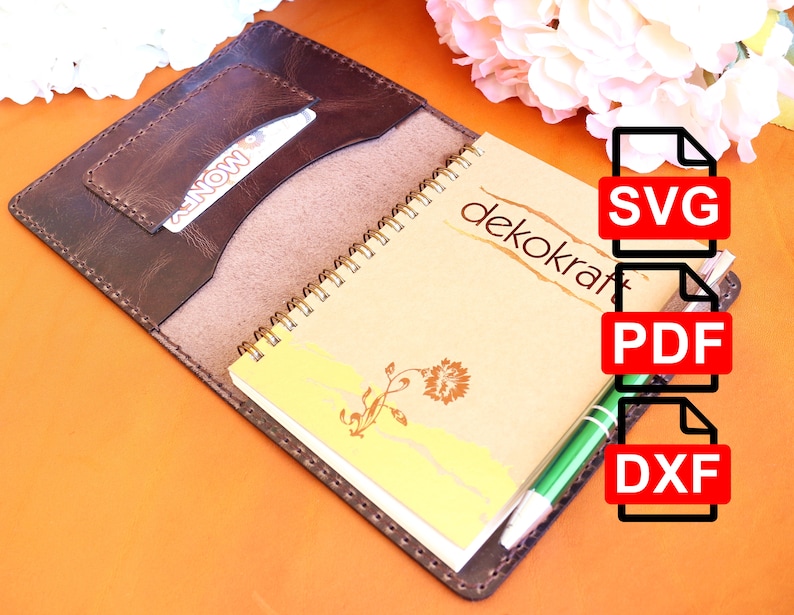 May include: A brown leather notebook cover with a pocket for a card or cash. The cover is open to reveal a spiral bound notebook with the word "dekokraft" printed on the front. The notebook is on a bright orange surface. The image also includes a green pen and three file icons labeled "SVG", "PDF", and "DXF".