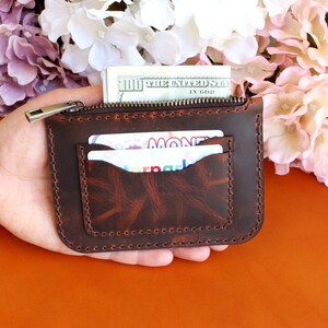 May include: A brown leather zippered wallet with two card slots and a pocket for cash. The wallet is held in a hand.