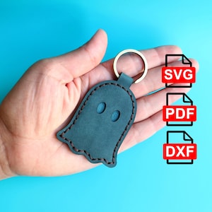May include: A black leather ghost keychain with a brown stitched outline. The keychain has a silver key ring and is held in a hand. The image also shows three red buttons with the text "SVG", "PDF", and "DXF" on them.