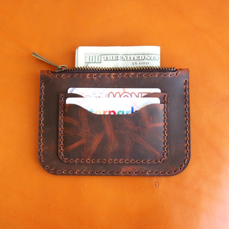 May include: A brown leather zippered wallet with a flap pocket and two card slots. The wallet is filled with cash and cards. "The United States of America" and "In God We Trust" are visible on the cash.