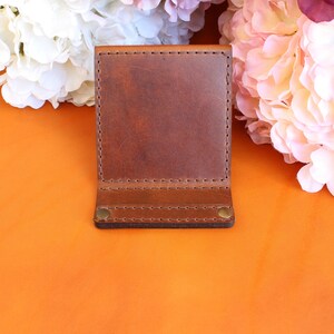 Puede incluir: Una cartera de cuero marr&oacute;n con un dise&ntilde;o de dos pliegues. La cartera est&aacute; hecha de cuero rico y texturizado y presenta costuras visibles a lo largo de los bordes. Dos remaches de lat&oacute;n aseguran la solapa inferior. El fondo incluye flores naranjas y rosas.