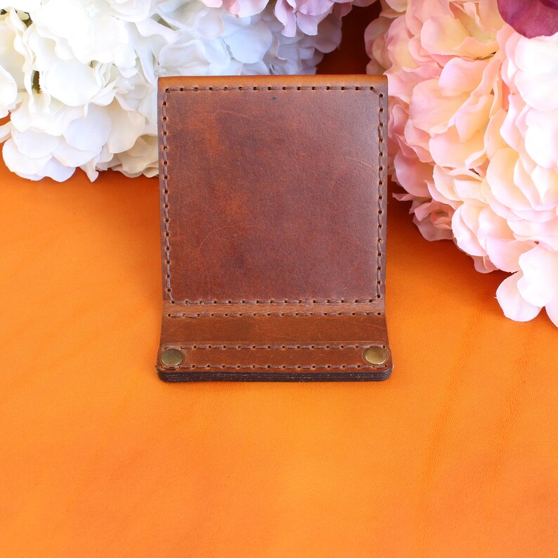 Puede incluir: Una cartera de cuero marr&oacute;n con un dise&ntilde;o de dos pliegues. La cartera est&aacute; hecha de cuero rico y texturizado y presenta costuras visibles a lo largo de los bordes. Dos remaches de lat&oacute;n aseguran la solapa inferior. El fondo incluye flores naranjas y rosas.