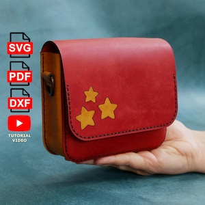 Puede incluir: Un bolso bandolera de cuero rojo con tres estrellas amarillas recortadas. El bolso tiene forma rectangular con cierre de solapa y un panel lateral marrón. El bolso se sostiene en una mano.