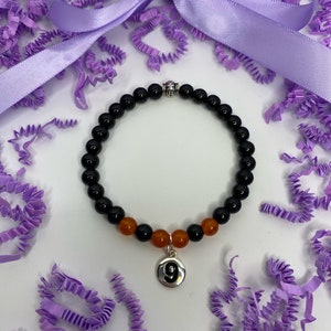 Puede incluir: Un brazalete de cuentas negras y naranjas con un dije plateado con el número 9. El brazalete se muestra sobre una superficie blanca con papel triturado morado.