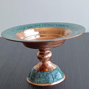 May include: A copper-coloured pedestal bowl with a turquoise speckled rim and base. The bowl has a wide, shallow dish and a rounded pedestal. The turquoise speckled design adds a decorative element to the bowl.