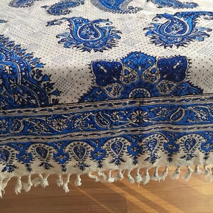 May include: A white and blue paisley patterned textile. The design features large, ornate paisley motifs in shades of blue against a white background. The border has a detailed blue pattern and white tassels. The textile is likely a tablecloth or decorative fabric.