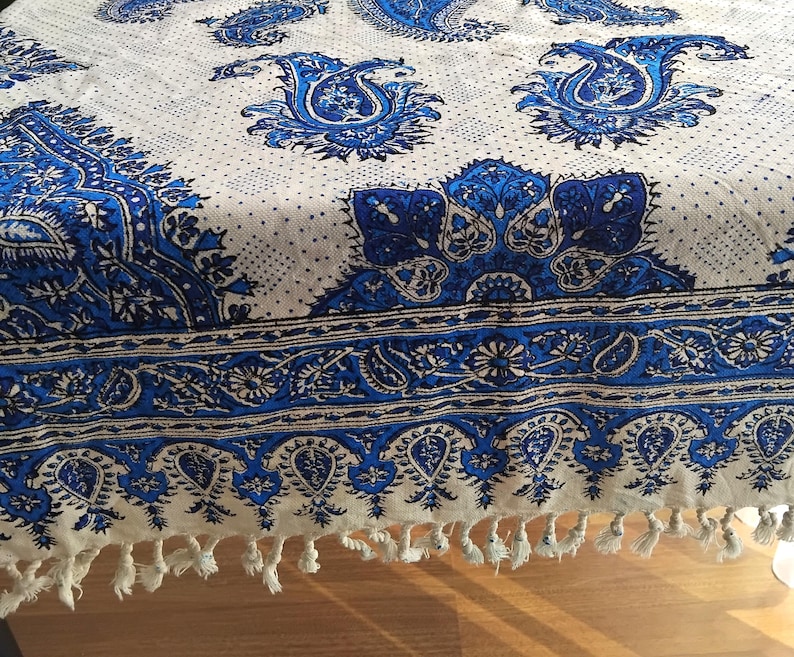 May include: A white and blue paisley patterned textile. The design features large, ornate paisley motifs in shades of blue against a white background. The border has a detailed blue pattern and white tassels. The textile is likely a tablecloth or decorative fabric.