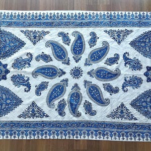 May include: A rectangular white and blue paisley patterned textile with a fringed edge. The design features large blue paisley motifs and decorative borders. The textile is likely a decorative throw or rug.