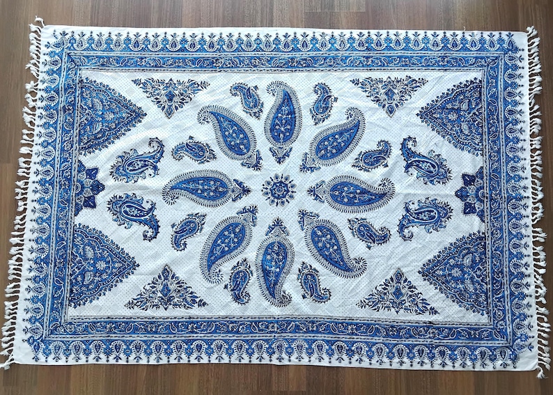 May include: A rectangular white and blue paisley patterned textile with a fringed edge. The design features large blue paisley motifs and decorative borders. The textile is likely a decorative throw or rug.