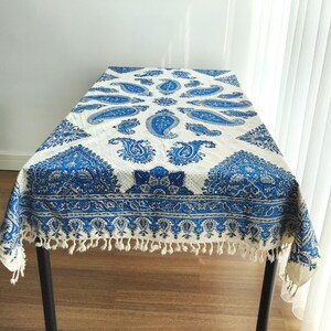 May include: A white and blue paisley patterned tablecloth. The tablecloth features a repeating paisley design in shades of blue against a white background. The edges are finished with a fringe.
