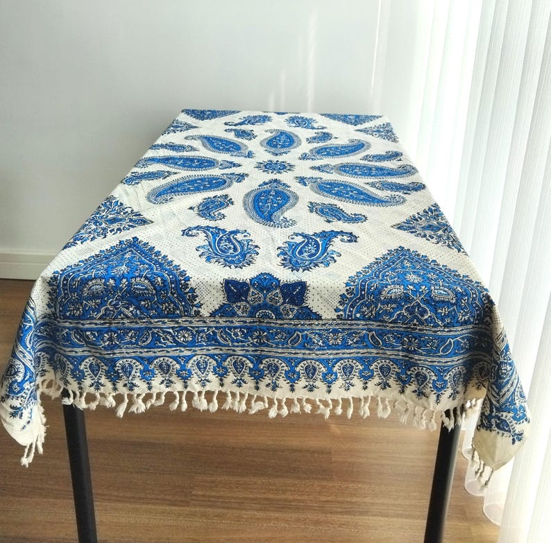 May include: A white and blue paisley patterned tablecloth. The tablecloth features a repeating paisley design in shades of blue against a white background. The edges are finished with a fringe.