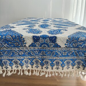 May include: A white and blue paisley patterned tablecloth. The tablecloth features a detailed design with blue paisley motifs and floral patterns. The edges are finished with a fringe. The tablecloth is on a wooden table.