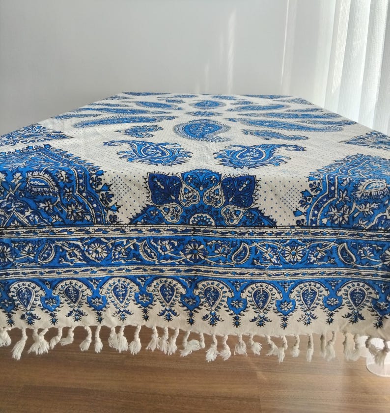 May include: A white and blue paisley patterned tablecloth. The tablecloth features a detailed design with blue paisley motifs and floral patterns. The edges are finished with a fringe. The tablecloth is on a wooden table.