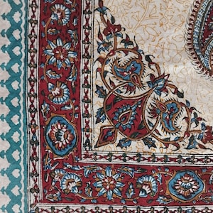 Può includere: Primo piano di un tessuto color crema con un design dettagliato a paisley e floreale. Il bordo presenta un motivo geometrico turchese. Il design centrale include elementi floreali e paisley rossi, blu e crema.