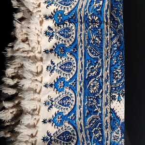 May include: Folded white and blue patterned textile with a paisley design. The textile features a border of cream-colored fringe. The design includes shades of blue and white, creating a decorative and textured appearance.