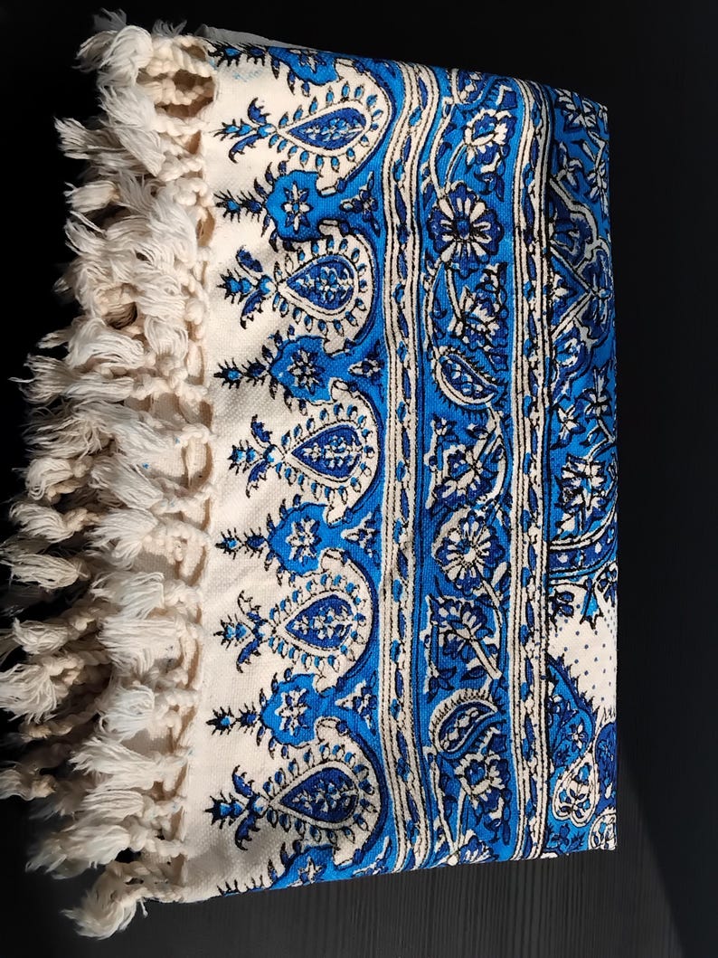 May include: Folded white and blue patterned textile with a paisley design. The textile features a border of cream-colored fringe. The design includes shades of blue and white, creating a decorative and textured appearance.