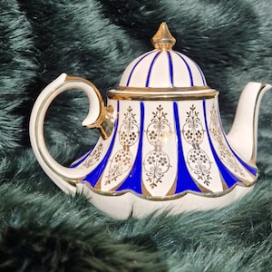 Op de afbeelding: Witte theepot met blauwe en gouden strepen en bloemmotieven. De theepot heeft een koepelvormig deksel met een gouden knop.