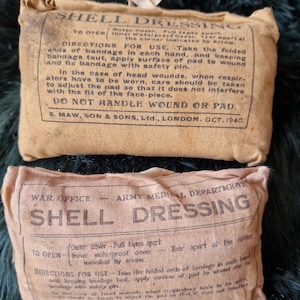 May include: Two vintage first aid kits, both labeled "Shell Dressing." The top kit has instructions for use, including how to open the kit and how to apply the bandage. The bottom kit has similar instructions, but also includes the text "War Office - Army Medical Department." Both kits are made of tan canvas.