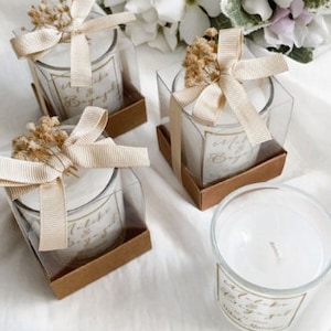 Puede incluir: Conjunto de cuatro pequeños tarros de velas de vidrio con tapas marrones y lazos de cinta beige. Cada tarro tiene una vela blanca con el texto "With love & best wishes" en letras doradas.