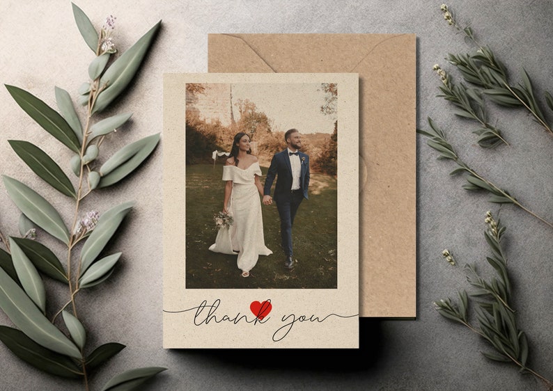 May include: A wedding thank you card with a photo of a bride and groom walking in a field. The card is beige with a red heart and the text "thank you" in cursive.