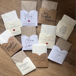 Peut inclure: Plusieurs petits paquets en papier, chacun contenant un mouchoir plié. Les paquets sont de différentes couleurs, avec du texte et des dates imprimés. Ils sont disposés sur une surface en bois.